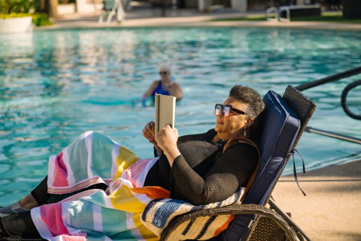 Resident enjoys reading by the pool while another exercises in the water, embracing wellness.