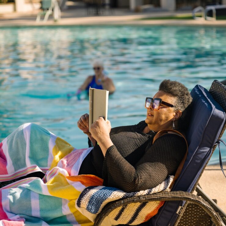 Resident enjoys reading by the pool while another exercises in the water, embracing wellness.