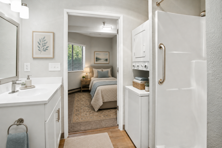 Bright bathroom with accessible shower, washer/dryer, and a comfortable bedroom visible beyond.