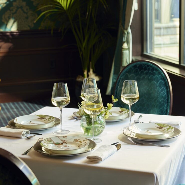 Sunlit dining table set for four with elegant place settings and plush teal chairs.