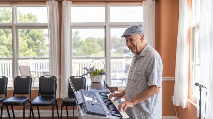 Senior man enjoying music on a keyboard in a bright, welcoming common room with large windows.