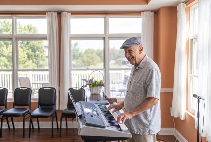 Senior man enjoying music on a keyboard in a bright, welcoming common room with large windows.