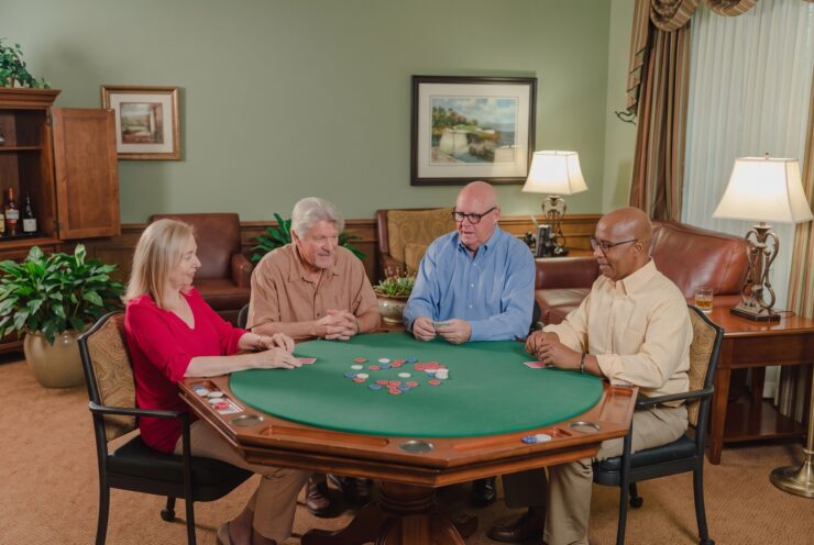 Four seniors enjoying a friendly card game in a warmly lit, inviting common room.