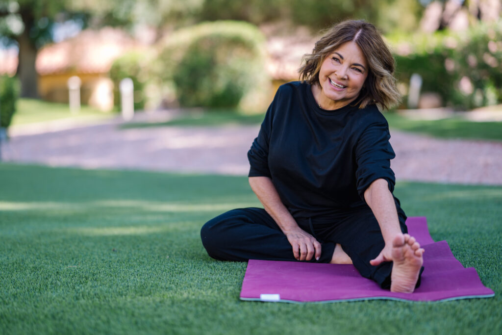 Woman smiling while stretching on a yoga mat outdoors in a peaceful garden setting