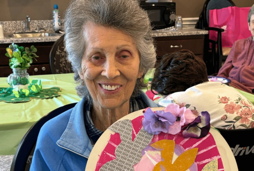 Elderly woman smiling warmly while showing a colorful craft project in a bright, welcoming room.
