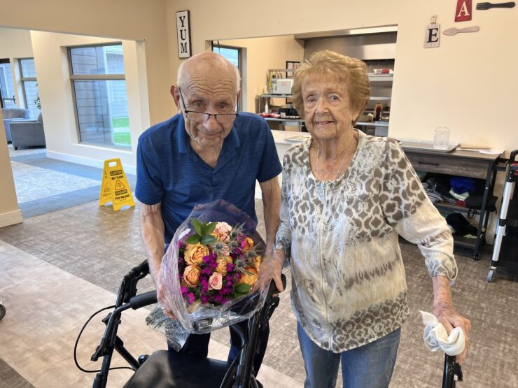 Two smiling seniors sharing a bright bouquet in a warm, inviting common area.