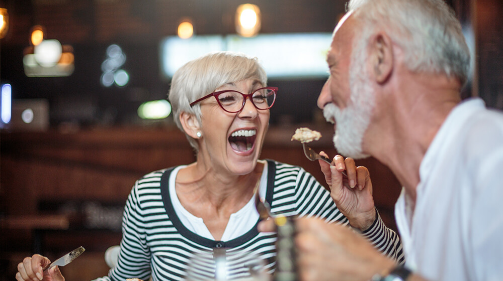 Older woman laughs and shares bite of her meal with husband in fancy restaurant