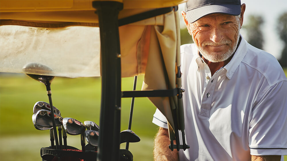 Senior gentleman cleans his golf club while standing beside golf cart on course
